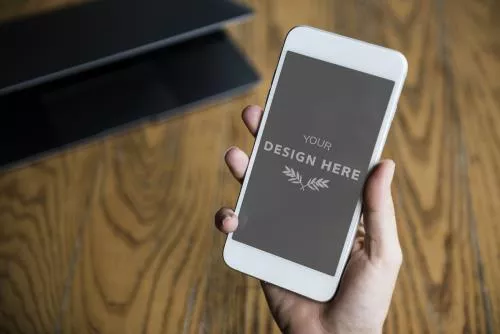 موکاپ موبایل سفید در دست یک آقا به پس زمینه میز چوبی و لپ‌تاپ فایل PSD قابل ویرایش 22057